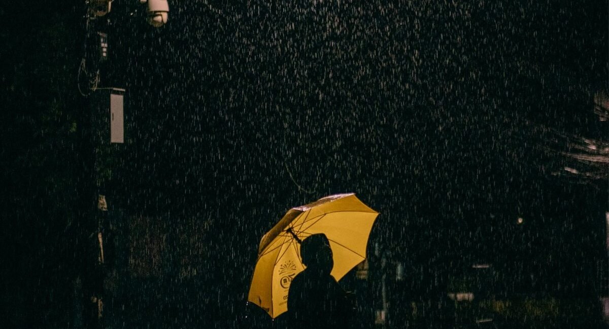 Pessoa com guarda-chuva amarelo sob chuva forte à noite, com as gotas iluminadas contra o fundo escuro