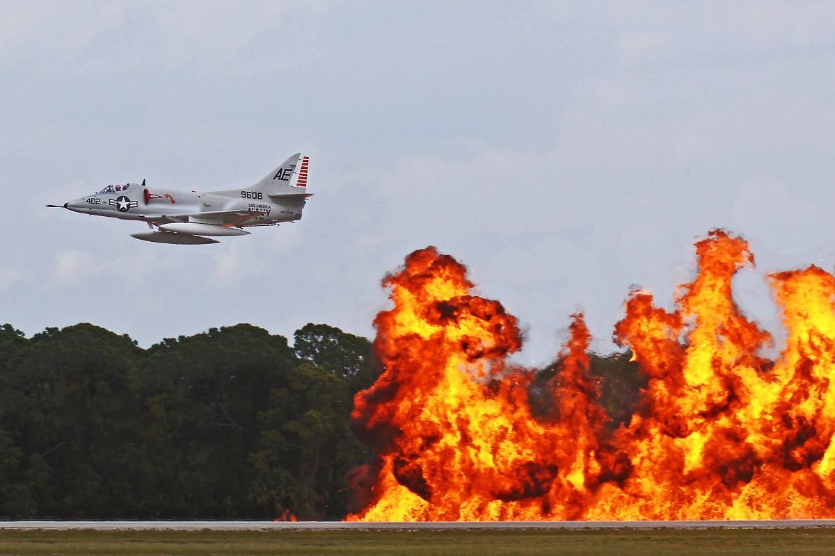 Avião a jato em voo rasante diante de uma parede de explosões pirotécnicas durante uma apresentação aérea