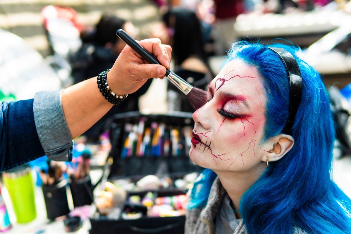 Maquiador aplica maquiagem artística com rachaduras vermelhas no rosto de uma mulher de cabelo azul