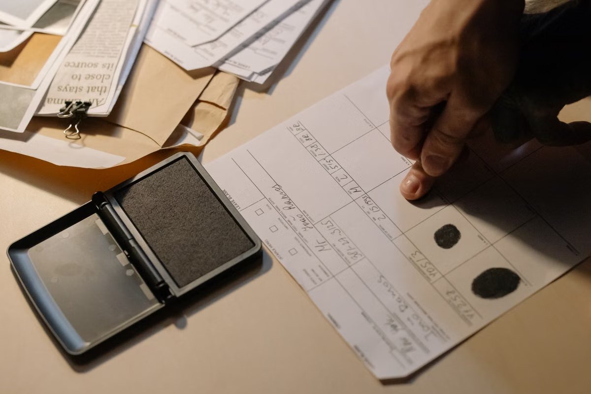 Mão registra impressões digitais numa ficha, ao lado de uma almofada de tinta e de pastas com documentos