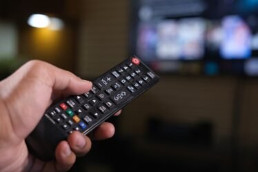 Mão segurando um controle remoto diante de uma TV desfocada